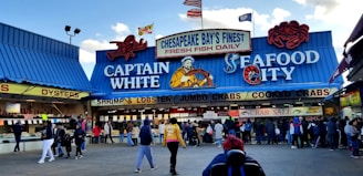 A bustling seafood market scene with a blue and red-themed storefront. The market features prominent signage advertising fresh fish, shrimp, lobster, jumbo crabs, and cooked crabs. Several people are gathered in front of the market, some seemingly in line or browsing.