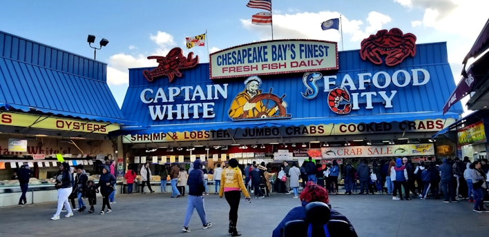 A bustling seafood market scene with a blue and red-themed storefront. The market features prominent signage advertising fresh fish, shrimp, lobster, jumbo crabs, and cooked crabs. Several people are gathered in front of the market, some seemingly in line or browsing.