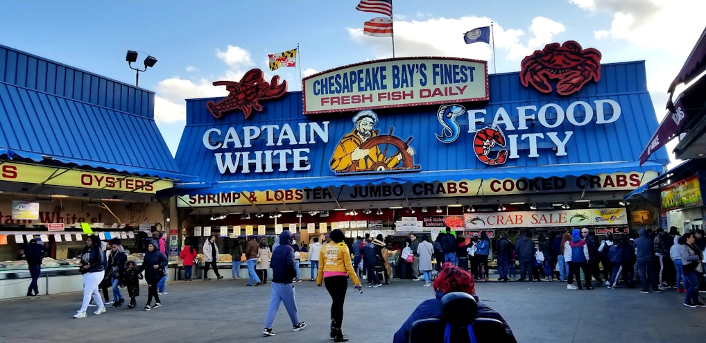 A bustling seafood market scene with a blue and red-themed storefront. The market features prominent signage advertising fresh fish, shrimp, lobster, jumbo crabs, and cooked crabs. Several people are gathered in front of the market, some seemingly in line or browsing.