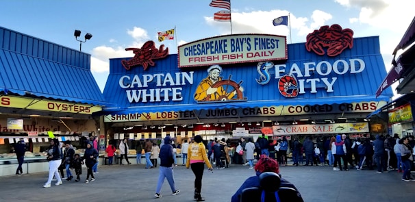 A bustling seafood market scene with a blue and red-themed storefront. The market features prominent signage advertising fresh fish, shrimp, lobster, jumbo crabs, and cooked crabs. Several people are gathered in front of the market, some seemingly in line or browsing.