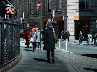 People are walking down a city sidewalk near a large building. A man in a black coat and beanie is walking away from the camera, carrying a green bag. A sign for a school of film and acting is visible. Other pedestrians are wearing winter coats, and the street appears busy with foot traffic.