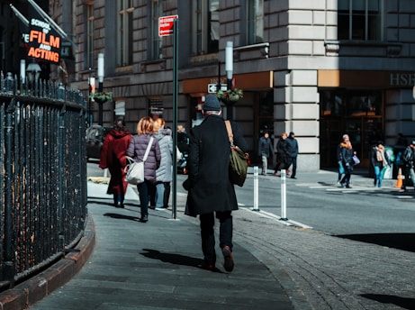People are walking down a city sidewalk near a large building. A man in a black coat and beanie is walking away from the camera, carrying a green bag. A sign for a school of film and acting is visible. Other pedestrians are wearing winter coats, and the street appears busy with foot traffic.
