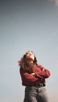 A woman with long curly hair stands confidently with her arms crossed, wearing a red sweater and jeans against a clear blue sky. Her face is lifted slightly upwards, suggesting a moment of tranquility and self-assurance.