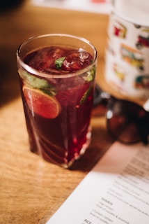 A glass filled with a reddish beverage containing ice cubes, mint leaves, and a slice of lime. The glass is placed on a wooden table, next to a menu and a pair of sunglasses. The light suggests a calm, relaxed atmosphere.