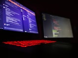 A dark room is lit by a laptop displaying two screens side by side. One screen displays a social media or messaging application with text conversations, while the other screen shows a text editor with what appears to be code. The laptop keyboard is illuminated with red backlighting.