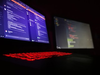 A dark room is lit by a laptop displaying two screens side by side. One screen displays a social media or messaging application with text conversations, while the other screen shows a text editor with what appears to be code. The laptop keyboard is illuminated with red backlighting.