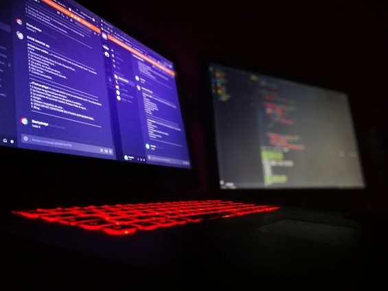 A dark room is lit by a laptop displaying two screens side by side. One screen displays a social media or messaging application with text conversations, while the other screen shows a text editor with what appears to be code. The laptop keyboard is illuminated with red backlighting.