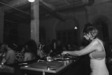 A black and white image depicts a DJ with headphones playing music on turntables in a dimly lit club. There is a crowd of people dancing and socializing in the background, highlighting a lively atmosphere. The setting is industrial, with exposed beams and pipes on the ceiling.
