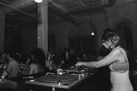 A black and white image depicts a DJ with headphones playing music on turntables in a dimly lit club. There is a crowd of people dancing and socializing in the background, highlighting a lively atmosphere. The setting is industrial, with exposed beams and pipes on the ceiling.