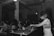 A black and white image depicts a DJ with headphones playing music on turntables in a dimly lit club. There is a crowd of people dancing and socializing in the background, highlighting a lively atmosphere. The setting is industrial, with exposed beams and pipes on the ceiling.