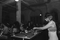 A black and white image depicts a DJ with headphones playing music on turntables in a dimly lit club. There is a crowd of people dancing and socializing in the background, highlighting a lively atmosphere. The setting is industrial, with exposed beams and pipes on the ceiling.