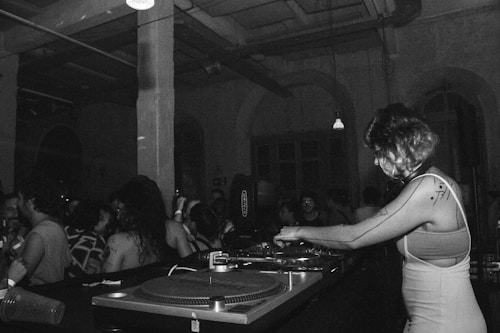 A black and white image depicts a DJ with headphones playing music on turntables in a dimly lit club. There is a crowd of people dancing and socializing in the background, highlighting a lively atmosphere. The setting is industrial, with exposed beams and pipes on the ceiling.