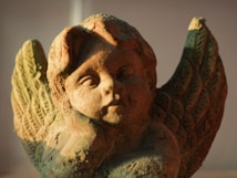 A close-up of a weathered stone cherub statue with detailed, intricately carved wings. The statue is illuminated by warm, soft lighting, casting subtle shadows that highlight its textured surface.