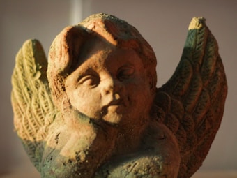 A close-up of a weathered stone cherub statue with detailed, intricately carved wings. The statue is illuminated by warm, soft lighting, casting subtle shadows that highlight its textured surface.