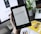 A person is holding a Kindle e-reader displaying text, with a laptop and plant in the background. The Kindle shows a page of text, which seems to be from a book. The background features a blurred computer screen and part of a magazine or book, creating a workspace ambience.