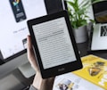 A person is holding a Kindle e-reader displaying text, with a laptop and plant in the background. The Kindle shows a page of text, which seems to be from a book. The background features a blurred computer screen and part of a magazine or book, creating a workspace ambience.