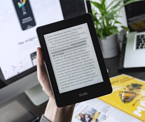 A person is holding a Kindle e-reader displaying text, with a laptop and plant in the background. The Kindle shows a page of text, which seems to be from a book. The background features a blurred computer screen and part of a magazine or book, creating a workspace ambience.