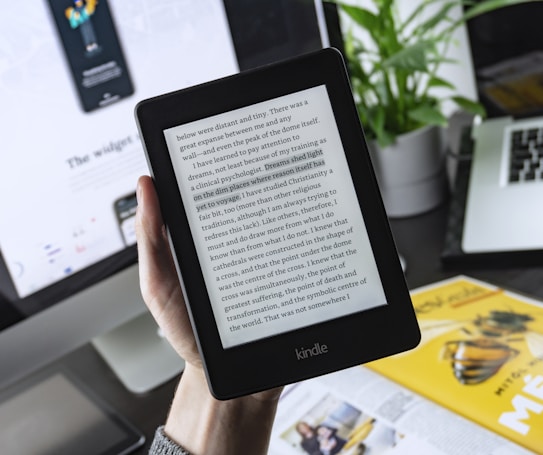 A person is holding a Kindle e-reader displaying text, with a laptop and plant in the background. The Kindle shows a page of text, which seems to be from a book. The background features a blurred computer screen and part of a magazine or book, creating a workspace ambience.