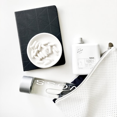 A minimalist arrangement featuring a black notebook with an embossed geometric pattern, a white dish filled with white capsules, a white square bottle labeled 'TONIK', a clear water bottle marked 'VOSS', and a white zippered bag with a subtle dotted pattern.