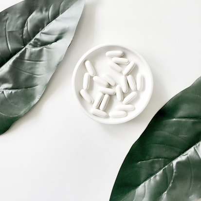 A white dish filled with white capsules is centered on a minimalist background. Two large, realistic-looking green leaves frame the dish on either side, creating a natural and clean aesthetic.
