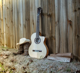 A wooden fence serves as the backdrop for an acoustic guitar resting on dry grass. To the left of the guitar, a pair of cowboy boots is placed, topped with a straw cowboy hat. On the right, several pieces of firewood are stacked on the ground. The scene evokes a rustic, country setting.
