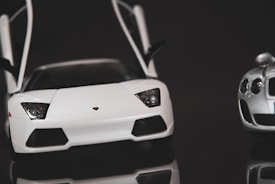 A close-up view of two sports cars with sleek designs. One car is primarily white with sharp, angular headlights and distinctive wing doors raised. The second car, mostly silver, is partially visible with round headlights. Both cars have a shiny finish and are set against a dark background, which enhances their glossy appearance.