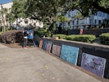 A person is painting on a canvas set on an easel in an outdoor setting surrounded by trees and bushes. Several completed artworks are displayed on the ground against a brick wall. The background reveals a white building with columns and a few people walking.