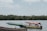 Several motorboats are floating on a calm body of water, with a lush green treeline in the background. One boat has a red canopy with text on it and appears to be carrying several people. Another boat is covered with a green tarp. The sky is cloudy but bright.