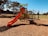 A children's playground with a red slide, climbing structures, and monkey bars stands on woodchip ground covering in a park setting. Trees and a lawn surround the play area under a clear blue sky, giving a sense of openness and leisure.
