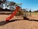 A children's playground with a red slide, climbing structures, and monkey bars stands on woodchip ground covering in a park setting. Trees and a lawn surround the play area under a clear blue sky, giving a sense of openness and leisure.