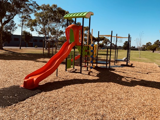 A children's playground with a red slide, climbing structures, and monkey bars stands on woodchip ground covering in a park setting. Trees and a lawn surround the play area under a clear blue sky, giving a sense of openness and leisure.