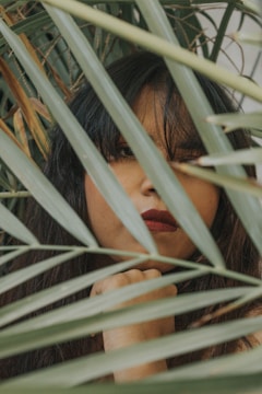 A person partially obscured by green palm fronds, visible with long dark hair and red lipstick, gazes directly at the camera. The setting feels intimate and slightly mysterious due to the natural frame formed by the leaves.