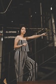 A woman stands on a staircase in a dimly lit environment, wearing a black and white striped outfit. Her posture is relaxed, with one hand holding the railing. The background features a dark metal structure and signs in Spanish.