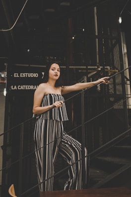 A woman stands on a staircase in a dimly lit environment, wearing a black and white striped outfit. Her posture is relaxed, with one hand holding the railing. The background features a dark metal structure and signs in Spanish.