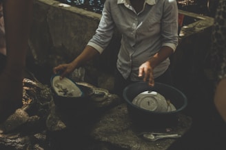 A person is washing dishes outdoors, using a plastic basin filled with water. The scene is shadowed and there is a rough stone surface beneath the basin. Several plates and utensils are visible.