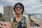 A person wearing a colorful tropical-patterned sleeveless top and large sunglasses is smiling broadly. In the background, there are tall buildings, a broad sandy beach, and the ocean under a cloudy sky. Red and white striped tape is visible in the foreground.