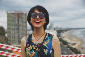 A person wearing a colorful tropical-patterned sleeveless top and large sunglasses is smiling broadly. In the background, there are tall buildings, a broad sandy beach, and the ocean under a cloudy sky. Red and white striped tape is visible in the foreground.