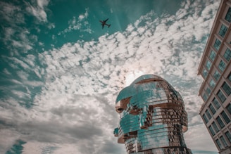 A modern sculpture made of reflective metallic elements is positioned against a backdrop of cloudy sky. An airplane is visible in the sky above, adding a sense of scale and motion to the scene. The architecture of a nearby building is partially visible on the right side, contributing to the urban environment.