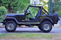 A black Jeep with an open top is parked on a driveway. The background features lush green foliage and a hint of a basketball hoop. The vehicle's shine contrasts with the natural surroundings, emphasizing its sturdy build designed for off-road use. The tires have visible branding and the vehicle appears well-maintained.
