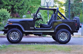 A black Jeep with an open top is parked on a driveway. The background features lush green foliage and a hint of a basketball hoop. The vehicle's shine contrasts with the natural surroundings, emphasizing its sturdy build designed for off-road use. The tires have visible branding and the vehicle appears well-maintained.