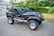 A black Jeep with an open-top design is parked on a driveway, featuring rugged tires and a well-maintained exterior. The vehicle appears ready for off-road use and is situated next to a suburban street with trees and houses in the background.