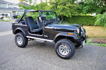 A black Jeep with an open-top design is parked on a driveway, featuring rugged tires and a well-maintained exterior. The vehicle appears ready for off-road use and is situated next to a suburban street with trees and houses in the background.