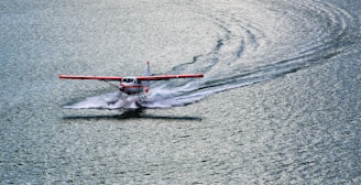 A seaplane with red wings is gliding across a large body of water, creating a wake behind it. The plane is positioned slightly to the left, and the water appears calm and reflective, with gentle ripples. The lighting suggests it's either early morning or late afternoon.