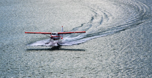 A seaplane with red wings is gliding across a large body of water, creating a wake behind it. The plane is positioned slightly to the left, and the water appears calm and reflective, with gentle ripples. The lighting suggests it's either early morning or late afternoon.