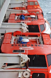 Several red lifeboats are lined up on the deck of a ship. The lifeboats are enclosed with large windows and equipped with release mechanisms that are visible. A railing and part of the ship's structure are adjacent to the lifeboats, and the ocean can be seen in the background.