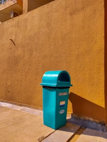 A teal-colored trash bin stands on a concrete pavement against a mustard-yellow textured wall. The bin is rectangular with a curved top and has informational stickers on it. The scene is brightly lit by sunlight, casting strong shadows.