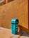 A teal-colored trash bin stands on a concrete pavement against a mustard-yellow textured wall. The bin is rectangular with a curved top and has informational stickers on it. The scene is brightly lit by sunlight, casting strong shadows.