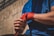 A person wearing a blue shirt is wrapping their hand with red boxing wraps, suggesting preparation for a physical activity, possibly boxing or martial arts. The background appears to be an outdoor space with a building wall.