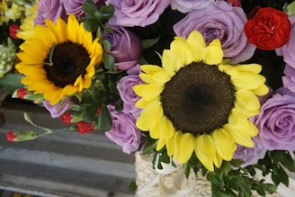 A vibrant arrangement of flowers featuring prominently two large sunflowers with bright yellow petals and rich brown centers. Surrounding them are soft purple roses and a scattering of small red flowers. The lush green leaves add freshness and depth to the composition.