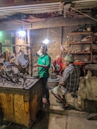 Several people are working in a dimly lit workshop. A man in a green shirt is standing near a table with various metal tools, another sits on a chair in plaid clothing, and a person in red takes a break sitting on a block in the background. The room contains worn brick walls, shelves cluttered with tools, and dim overhead lighting.
